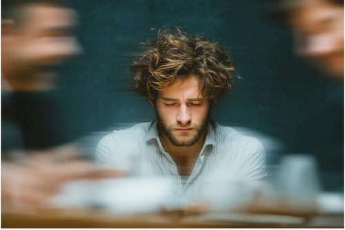 A man appearing stressed and overwhelmed while working in a busy café, surrounded by blurred figures in the background